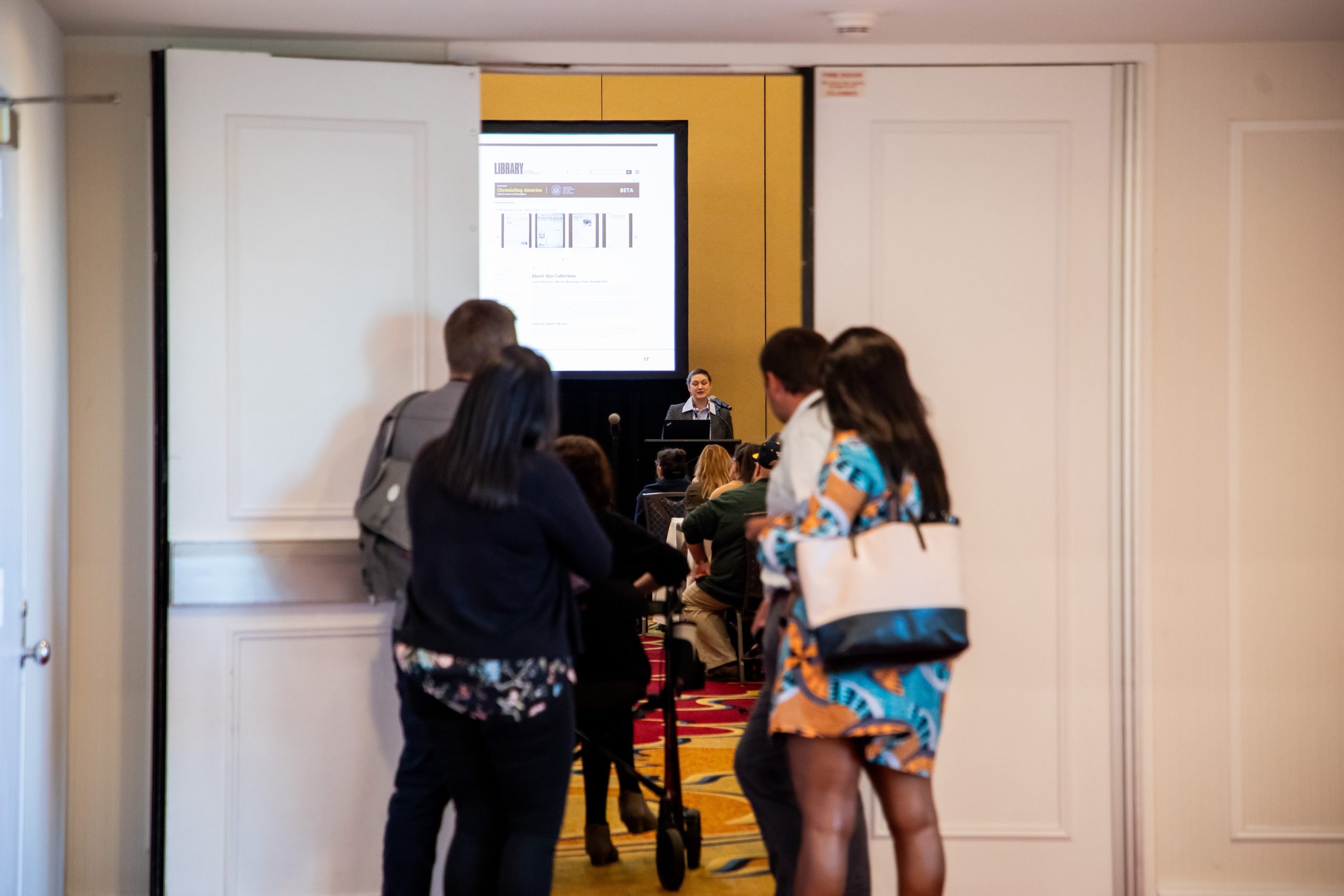 People standing at doorway of a conference session room, also showing presenter at podium with slideshow in background