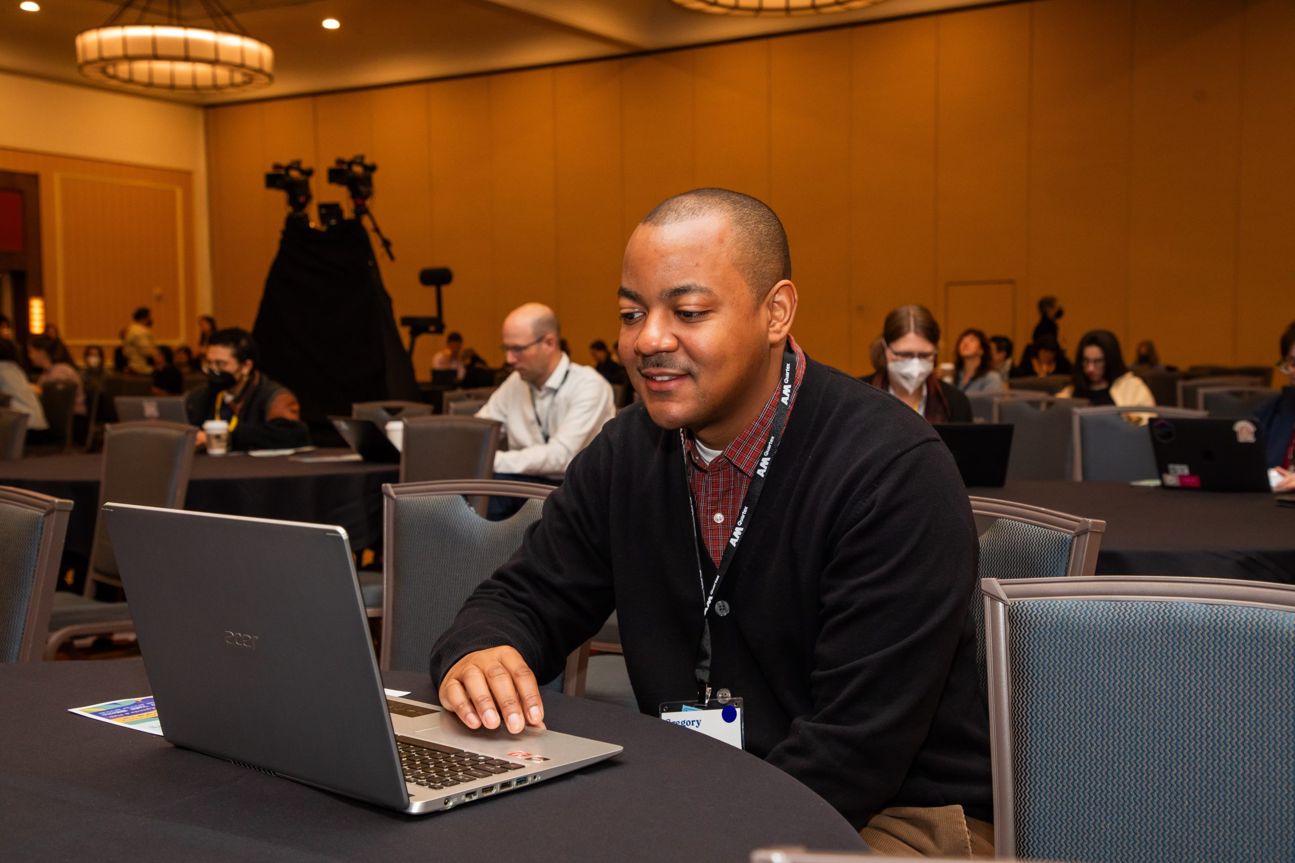 Person seated at a table on a laptop with people in the background.