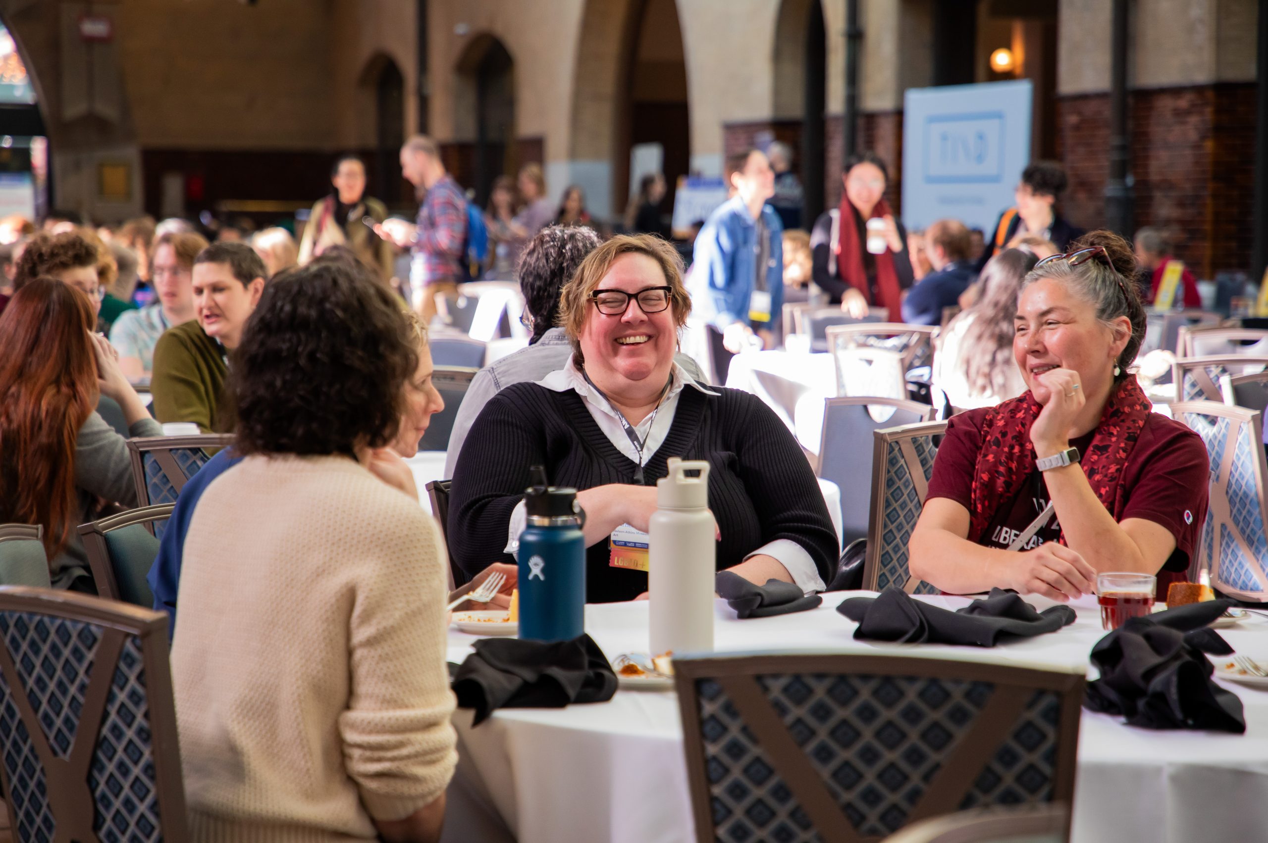 People seated in a conference dining/exhibition area. Three people seated at a round table having a conversation in the foreground.