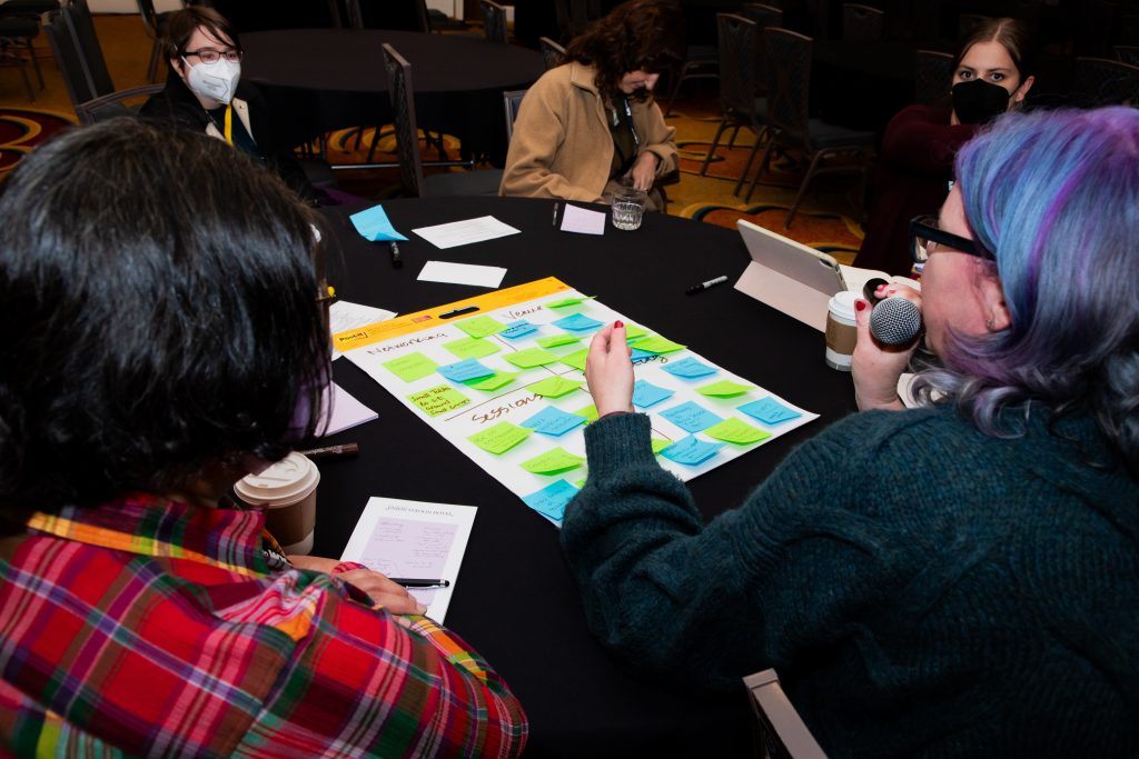 View of people at a table from behind, showing a person with purple hair holding a microphone with large sticky note and smaller sticky notes on table.