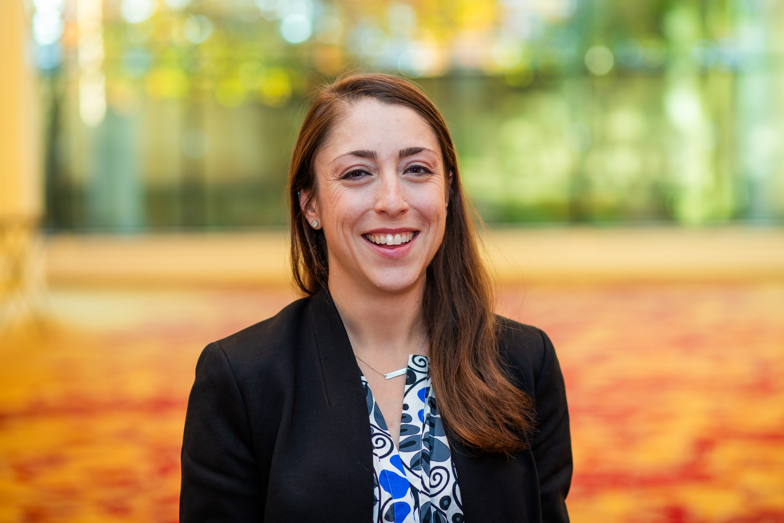 Aliya Reich headshot - Aliya wearing a blazer with a blurred hallway in the background