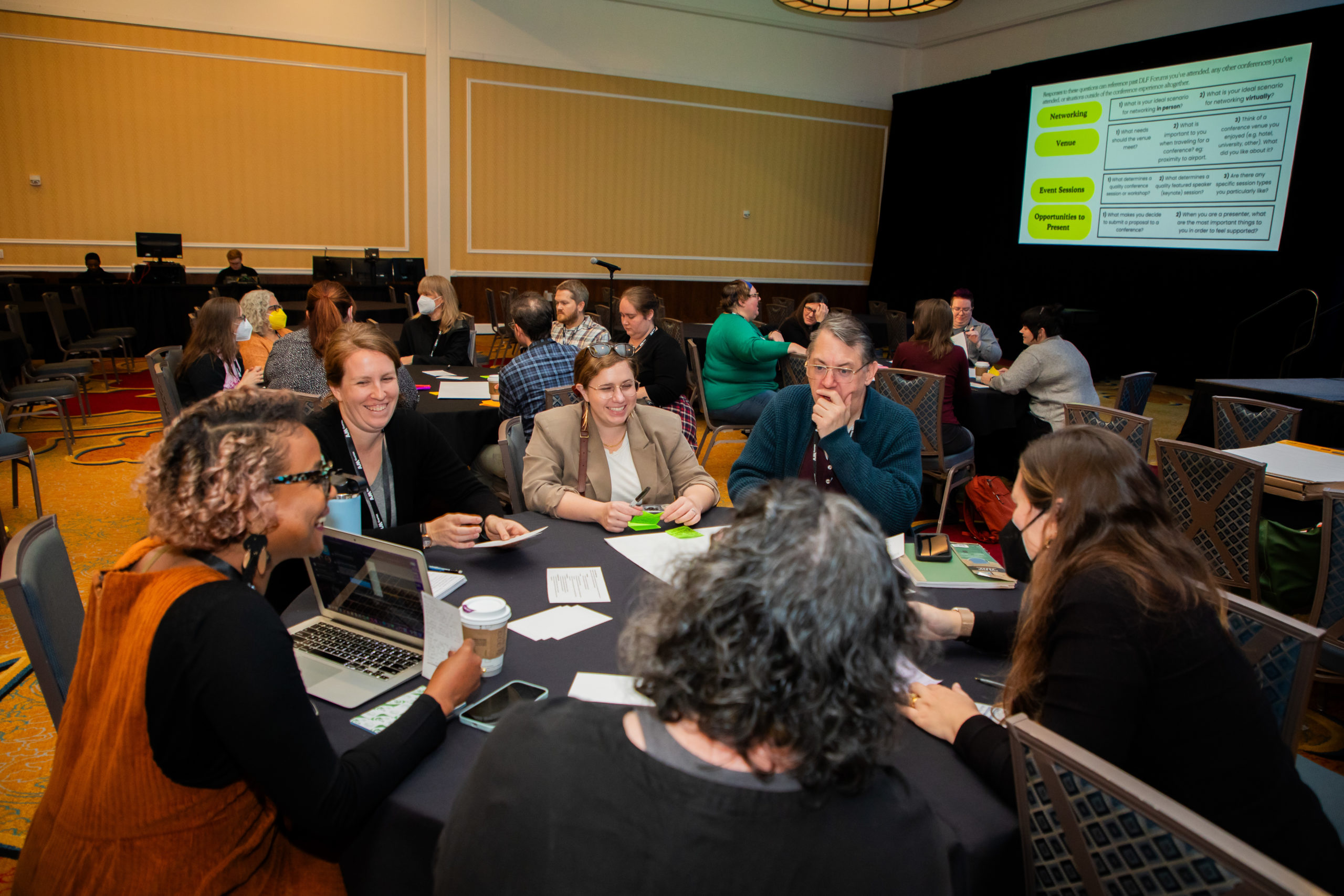 People seated at tables in a conference hotel main space with projector screen in background.