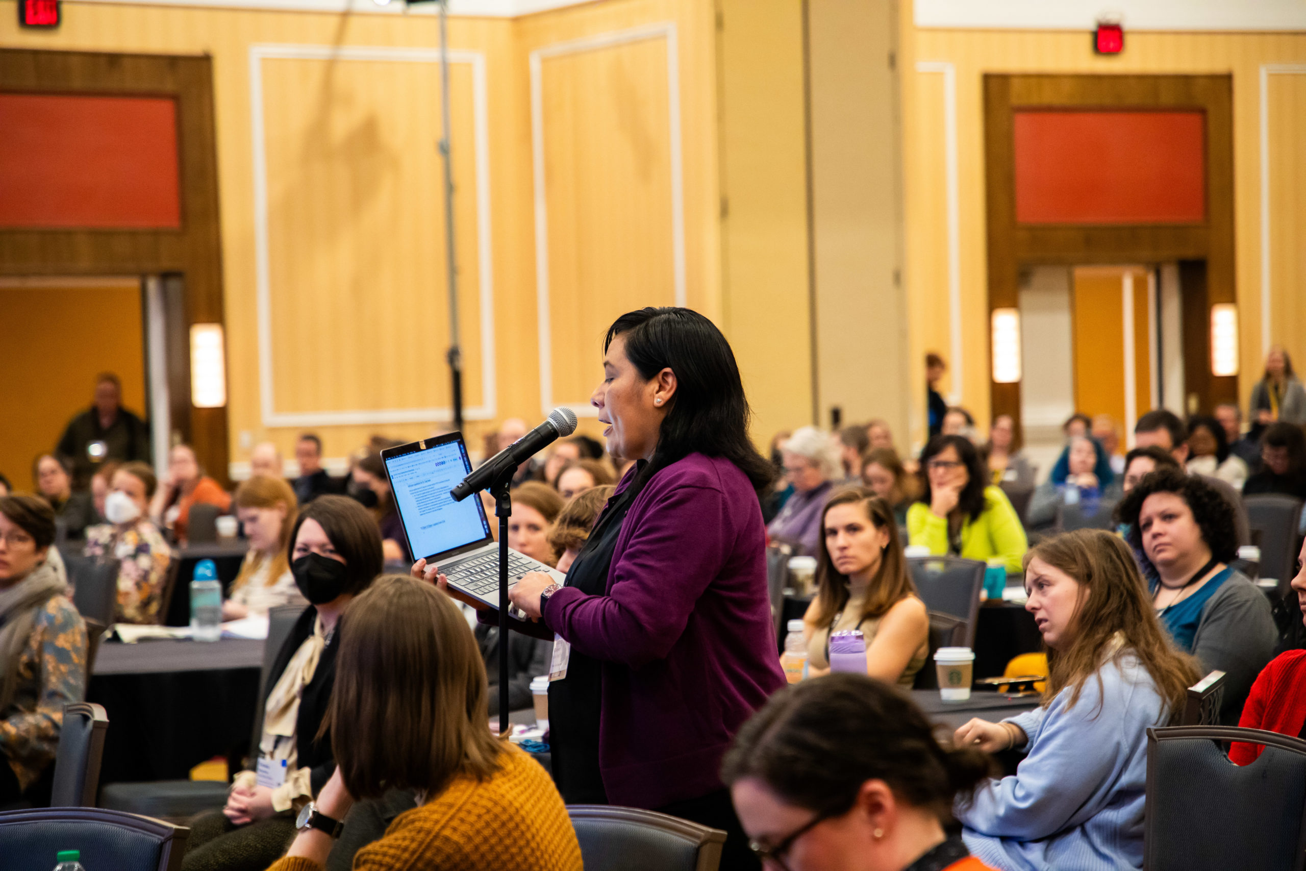 Person holding a laptop at a microphone in a hotel conference room with people seated at tables.